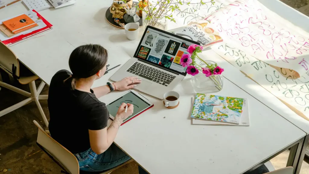 The future of remote work: A young professional works from a creative home office using a laptop and tablet, surrounded by design materials and coffee, showcasing flexibility and digital collaboration.