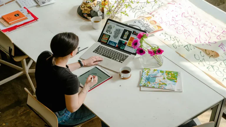 The future of remote work: A young professional works from a creative home office using a laptop and tablet, surrounded by design materials and coffee, showcasing flexibility and digital collaboration.