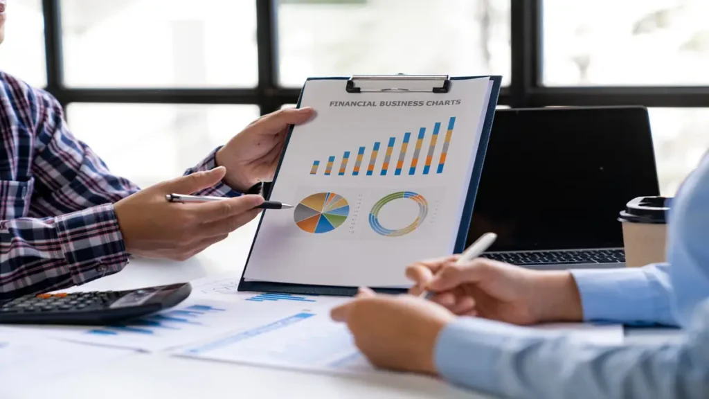 Two professionals discussing financial business charts with graphs and data on a clipboard during a meeting